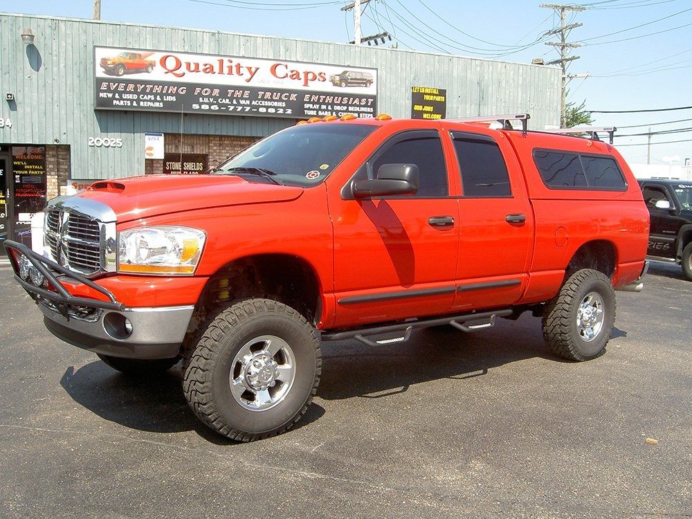A red Dodge pickup truck with a camper shell parked in front of a Quality Caps shop.