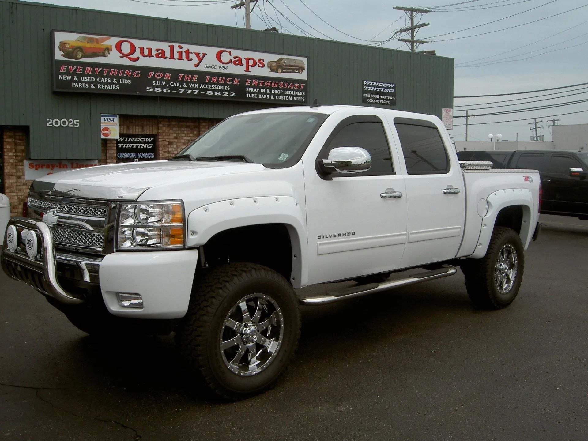 A white pickup truck with a chrome grille and lifted suspension parked in front of a Quality Caps storefront.