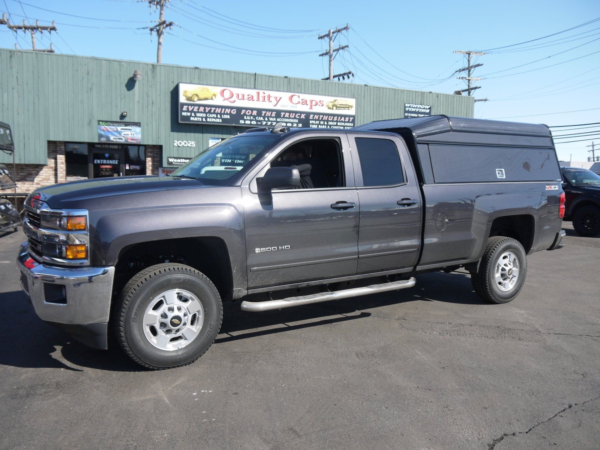 A dark gray Chevrolet pickup truck with a matching camper shell parked in front of a Quality Caps storefront.