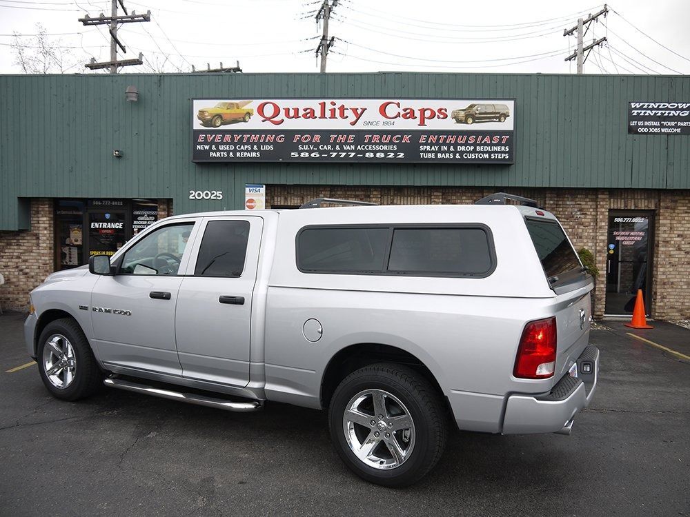 A silver pickup truck with a matching camper shell parked in front of a storefront sign reading Quality Caps.