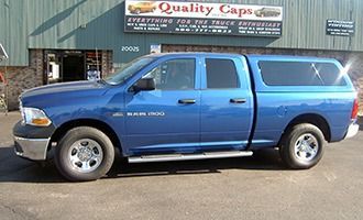 A bright blue Ram 1500 truck with a matching camper shell parked in front of a shop named Quality Caps.