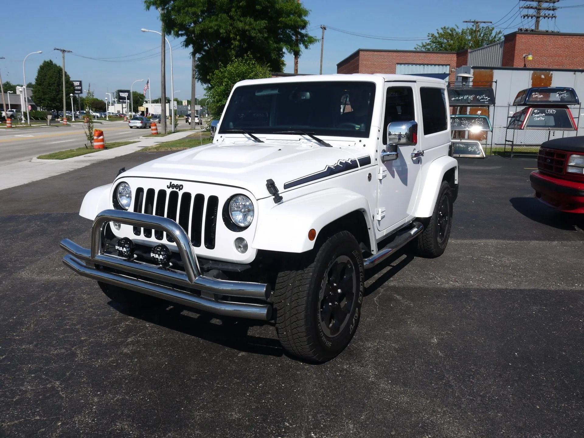A white two-door Jeep Wrangler with a chrome grille guard parked in an outdoor lot on a sunny day.