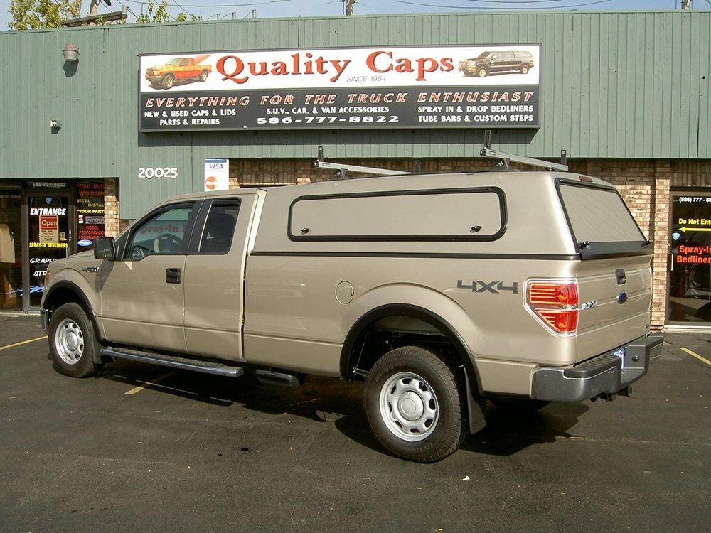 A tan 4x4 pickup truck with a camper shell parked in front of the Quality Caps store.
