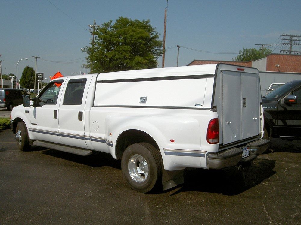 A white dually pickup truck with a white utility cap parked in a sunny car lot.