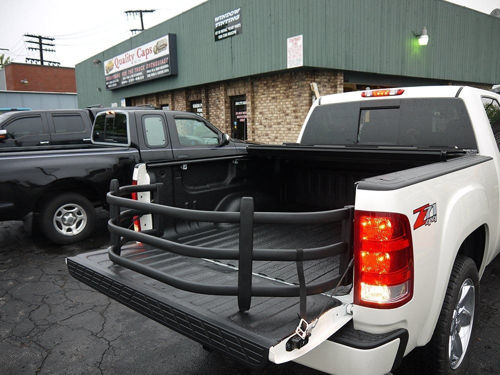 A white pickup truck with a black cargo bed extender parked in front of a brick and green business building.