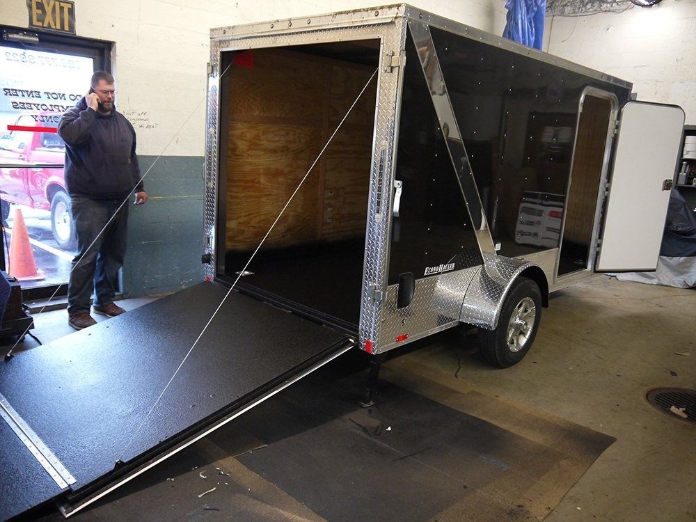 A black enclosed cargo trailer with a lowered ramp door sits inside a warehouse, with a person standing by the entrance.