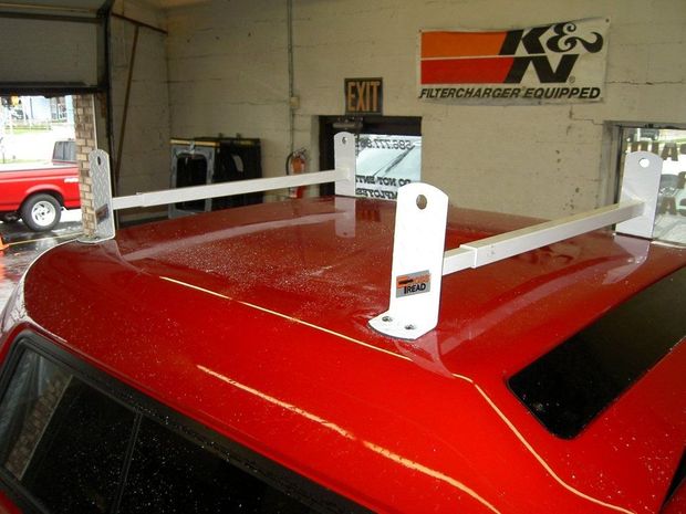 White metal utility ladder racks installed on the red roof of a pickup truck camper shell inside a garage.