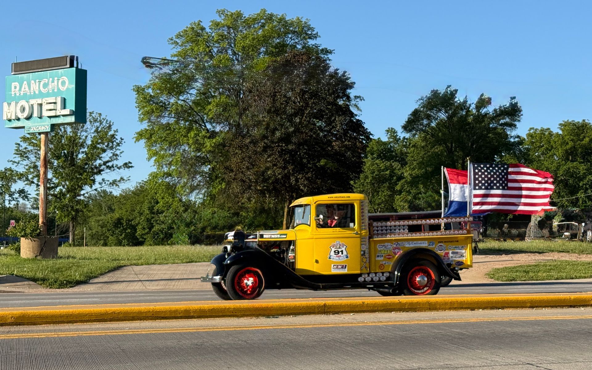 Yellow vintage race truck drifting on a track, kicking up dust, with American and blue flags waving overhead