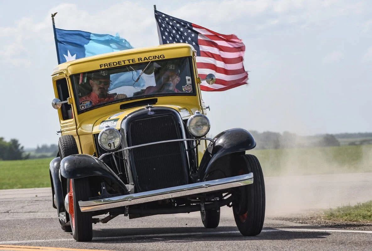 Yellow vintage race car drifting on a track, kicking up dust, with American and blue flags flying behind it