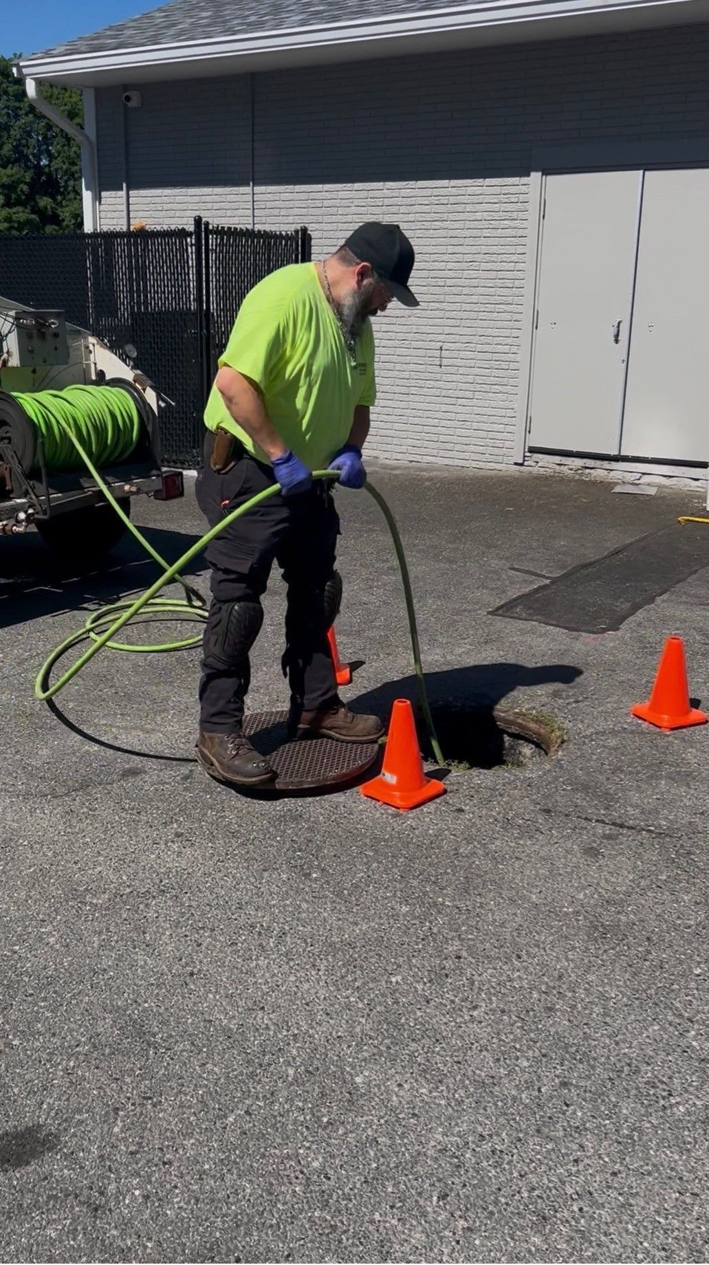 Worker in neon shirt using a hose to clean a pothole in a parking lot, with orange cones nearby.