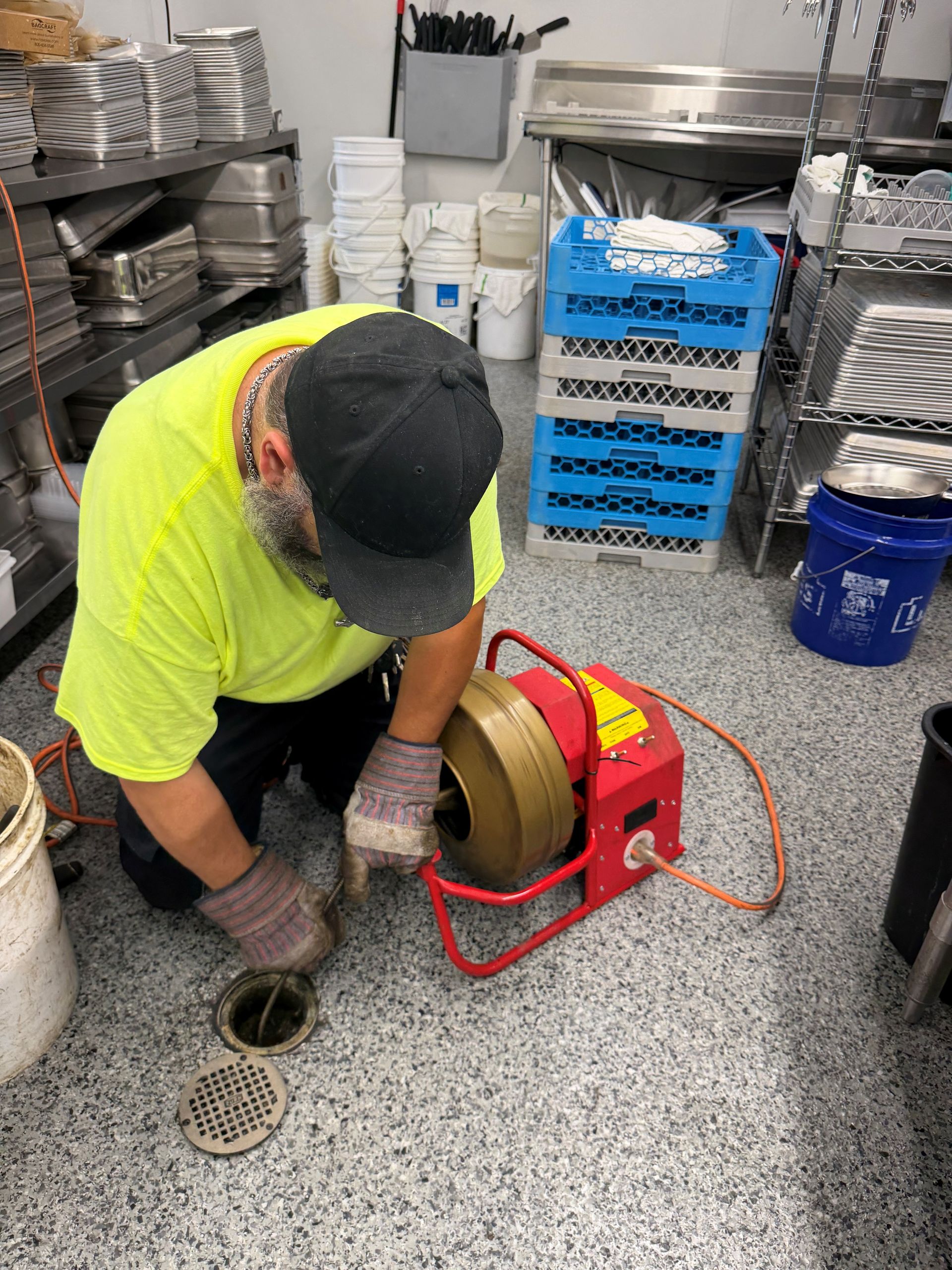 Worker in neon shirt using a drain auger beside floor drains in a commercial kitchen.