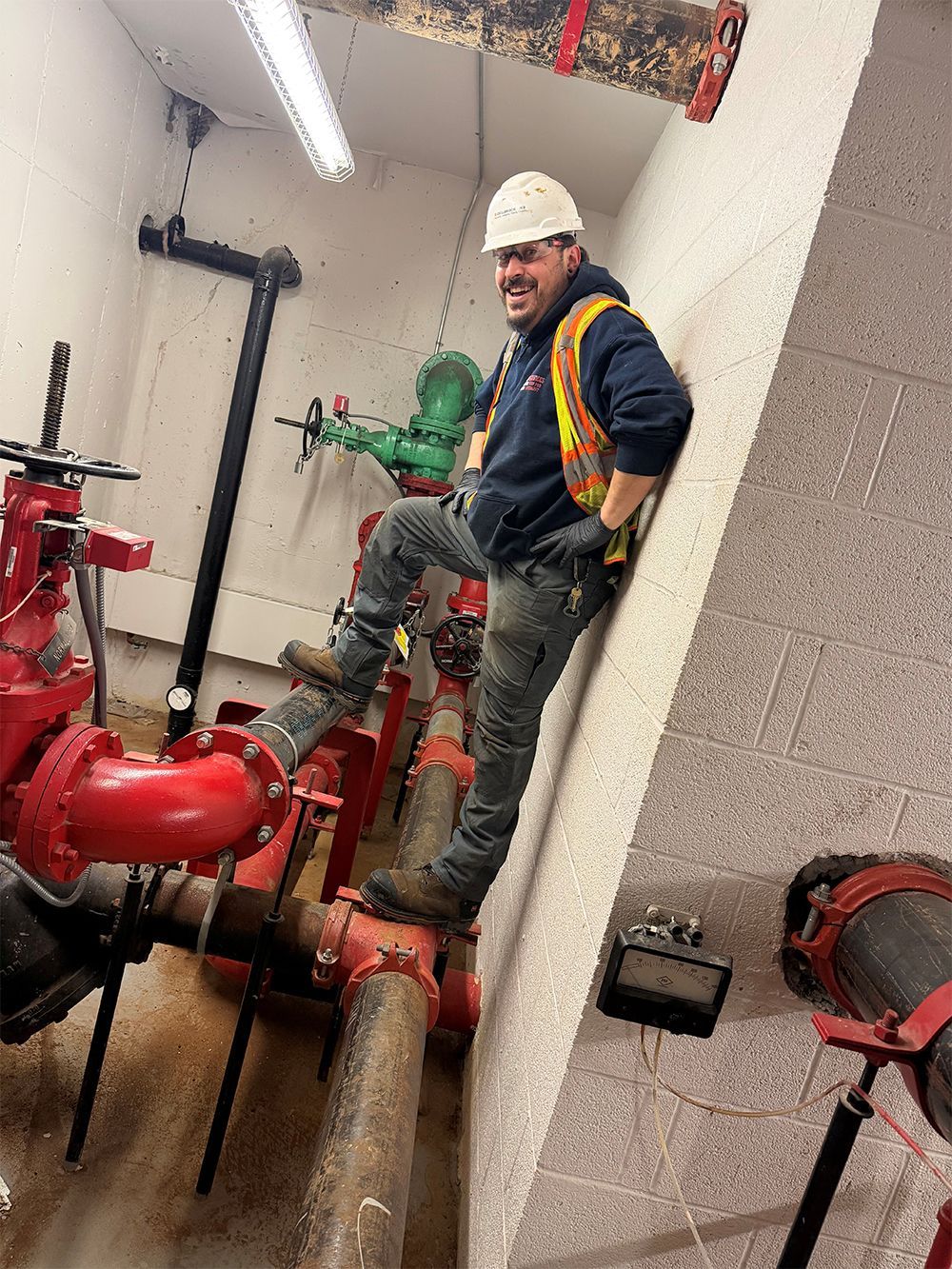 A smiling worker in safety gear, hard hat, and high-visibility vest stands with one foot on a large industrial pipe.