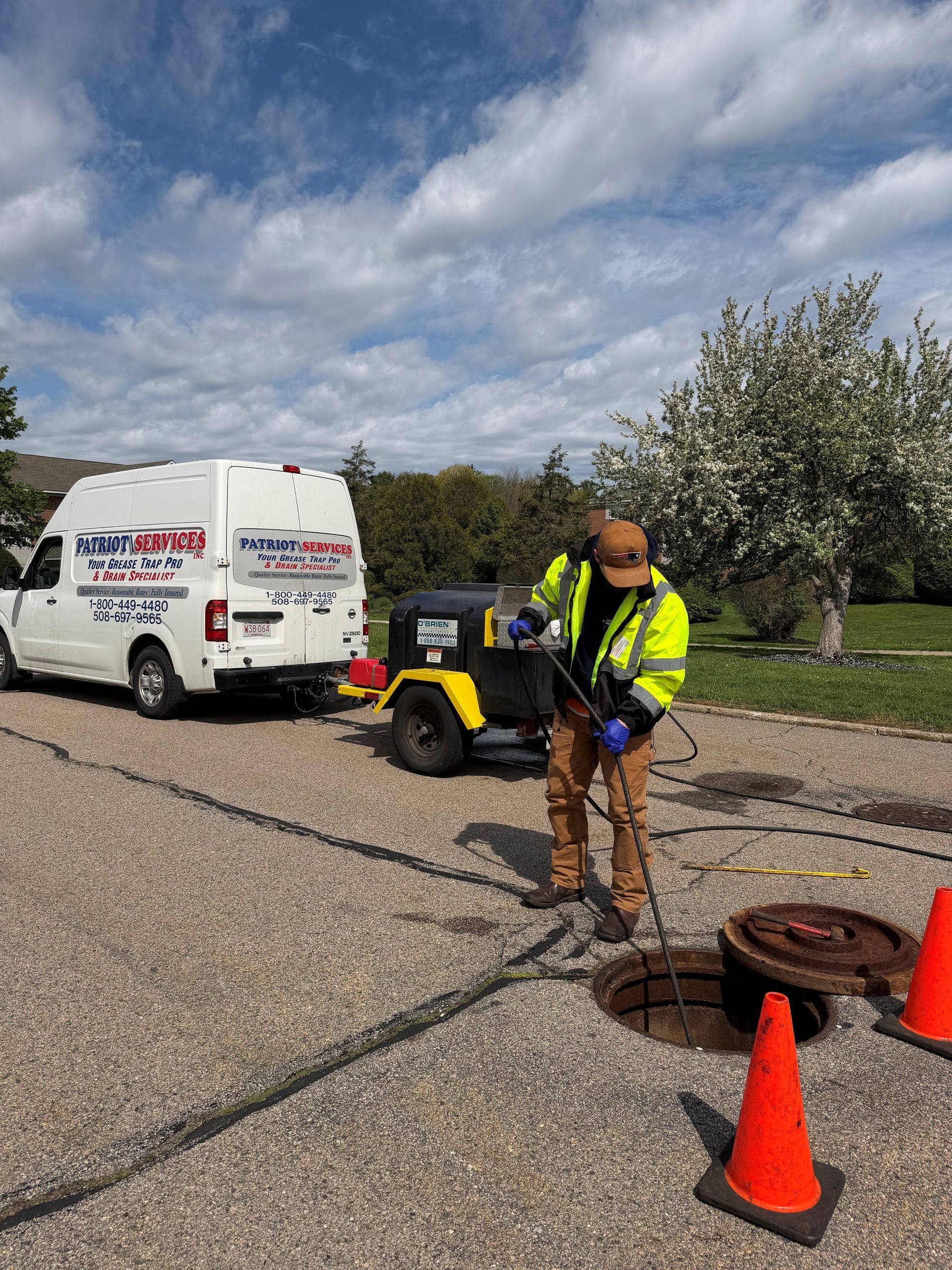 Worker in neon jacket clears a road drain beside a utility van and traffic cones.