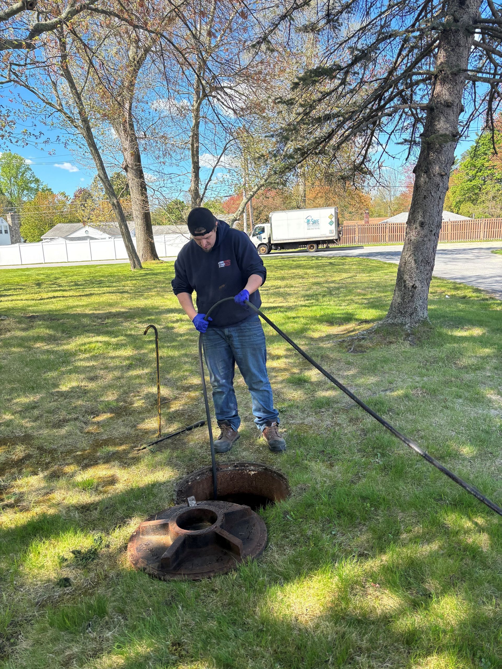 Person using a long tool near an open ground hole in a grassy park with trees nearby