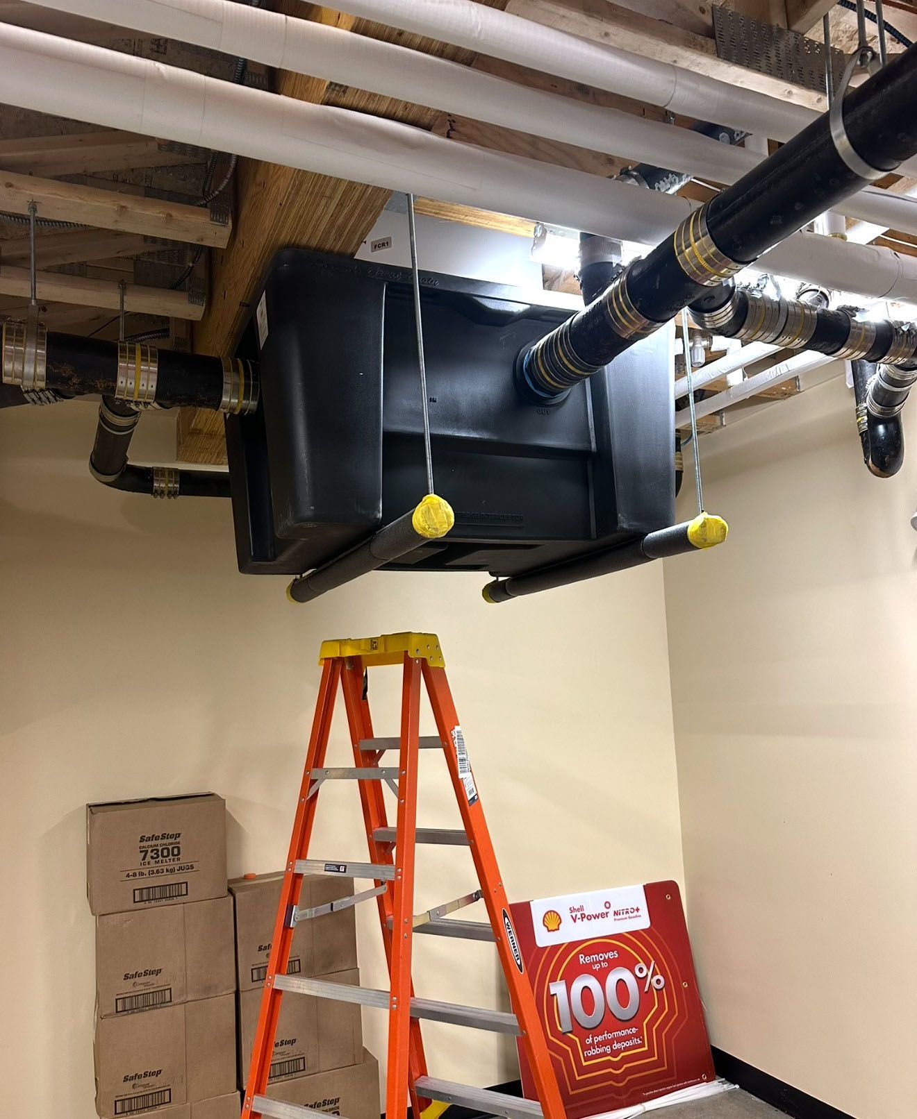 A black tank suspended from ceiling pipes with a ladder below. Yellow accents and cardboard boxes are also present.