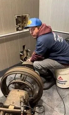 Man in blue hat repairs machinery near a wall. He sits on a bucket.