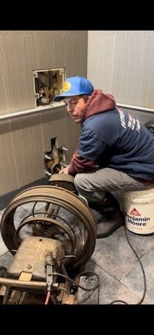 A worker repairs equipment near a wall. He kneels beside a large appliance. A paint bucket sits nearby.