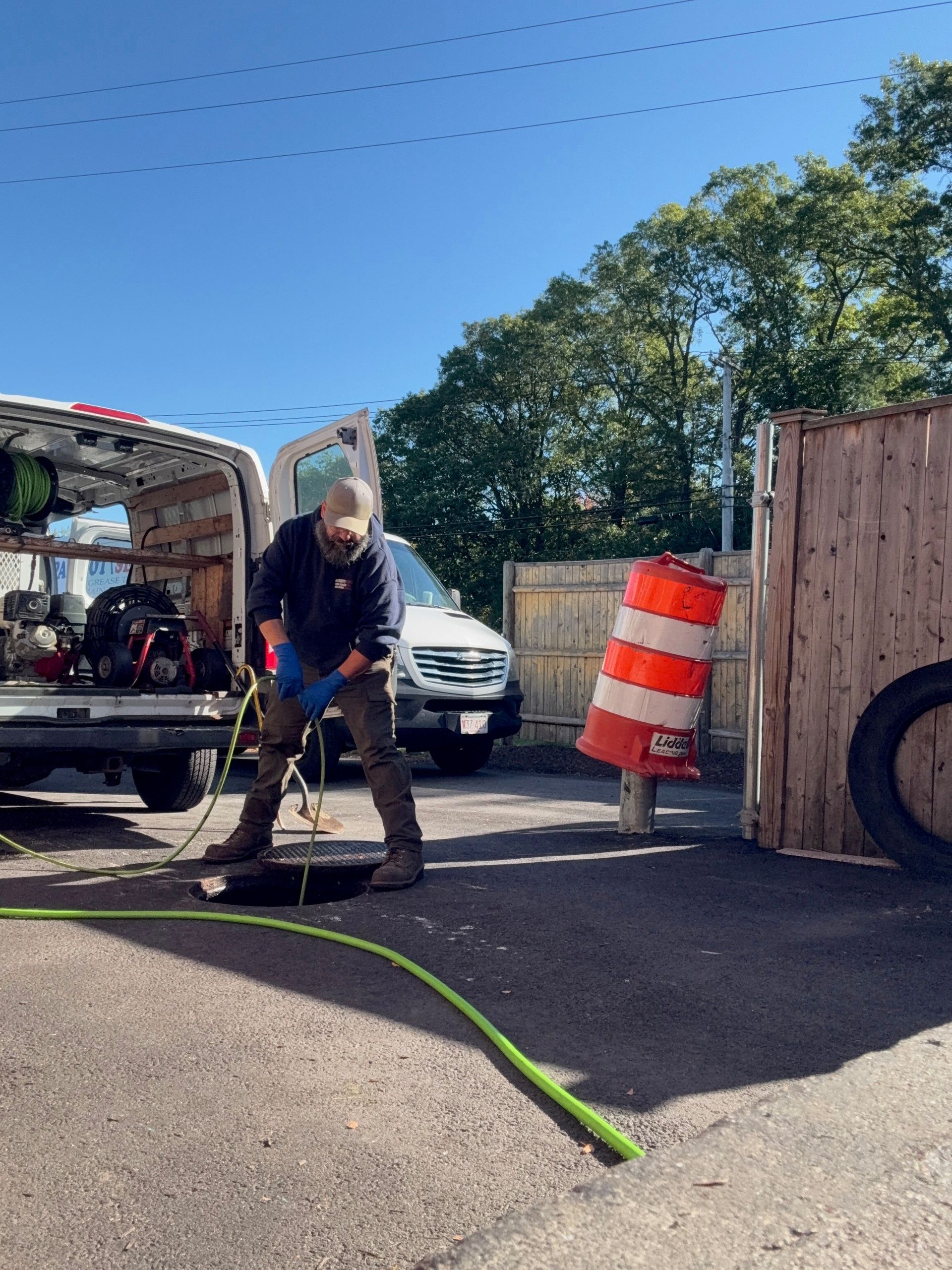 A man in a blue shirt clearing a sewer drain near a van and traffic barrel.