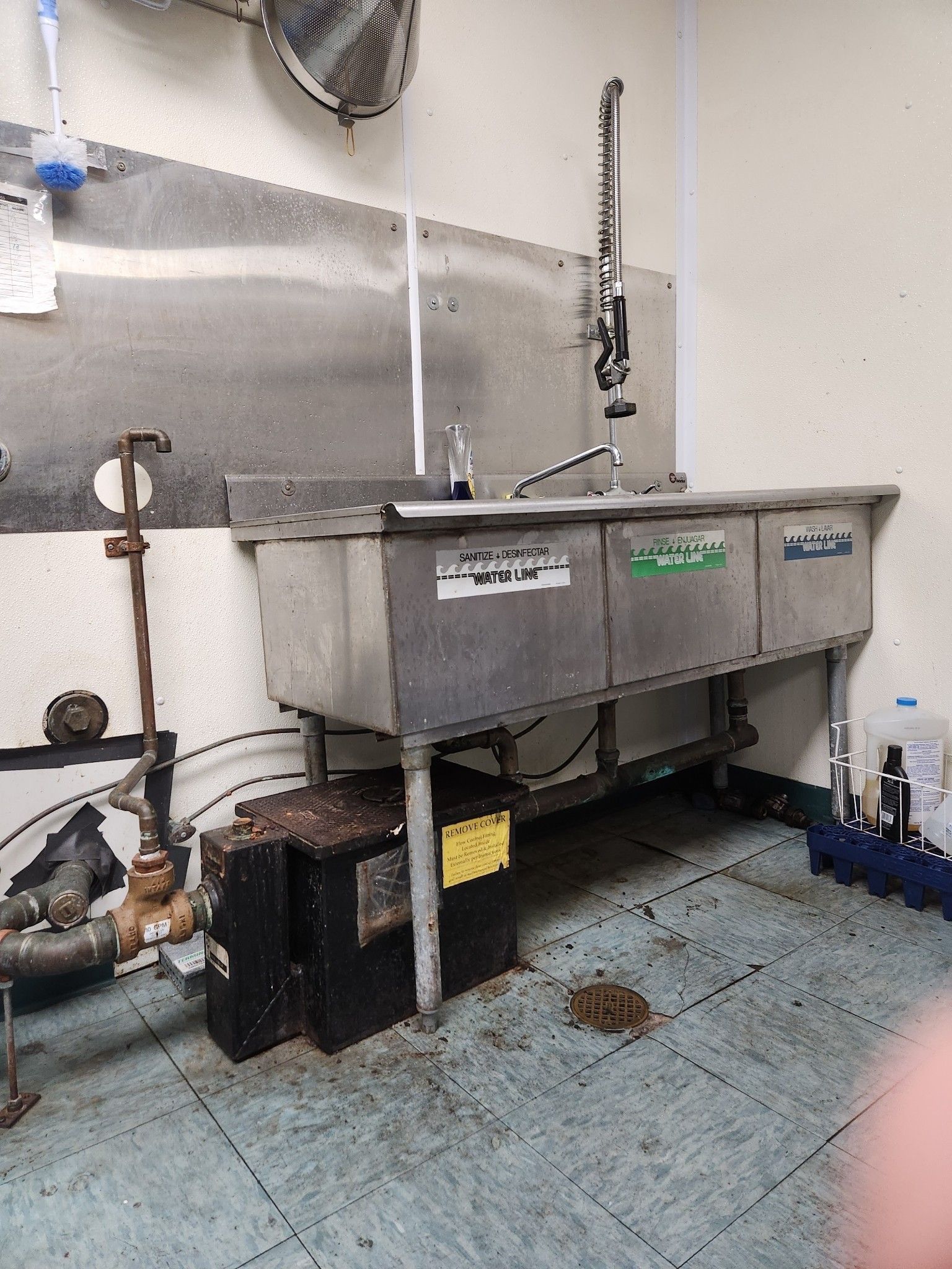 Stainless steel commercial sinks in a kitchen, with a dirty floor and pipes.