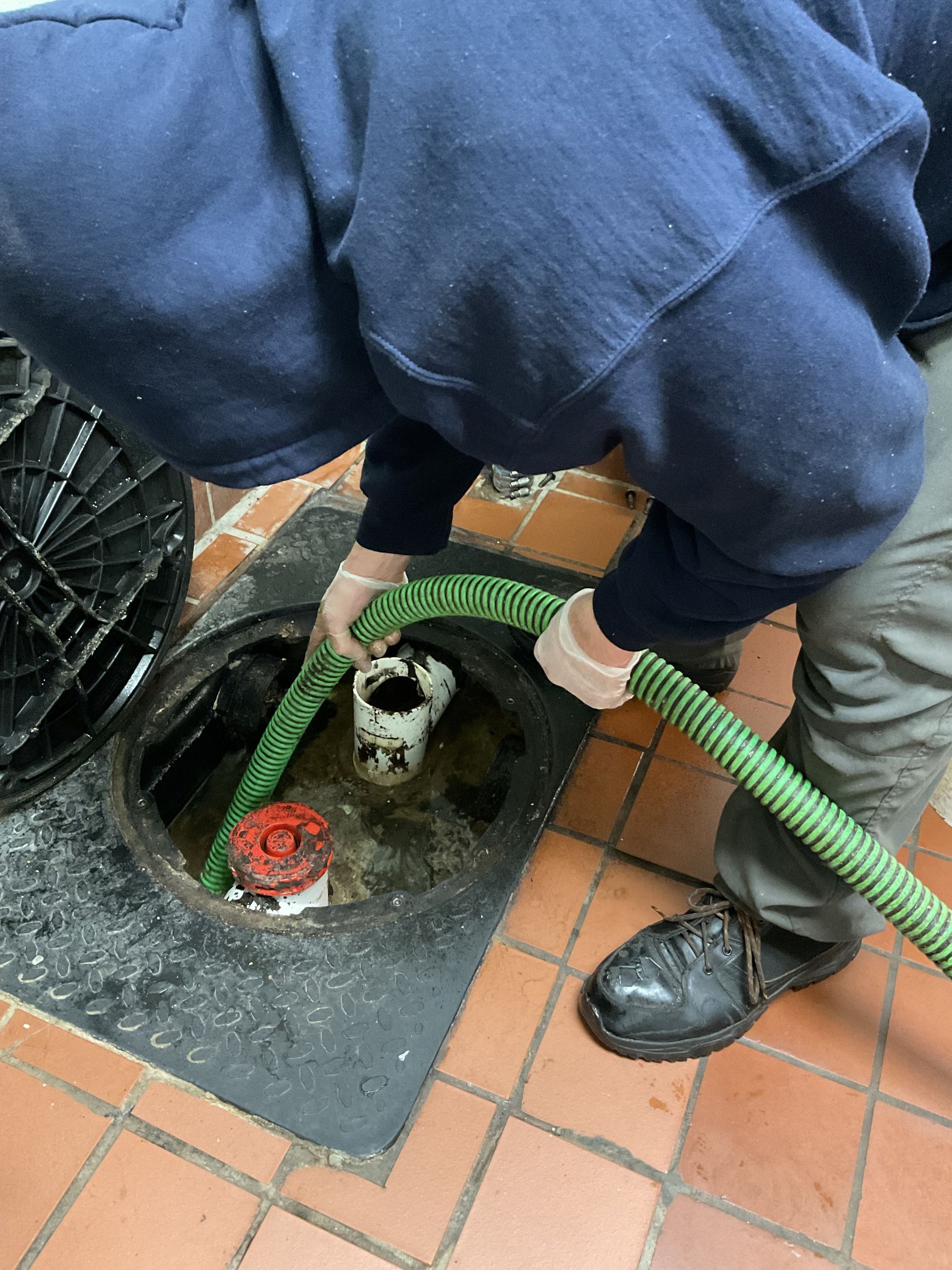 Person in blue sweater and gloves pumping liquid from a grease trap in a commercial kitchen.