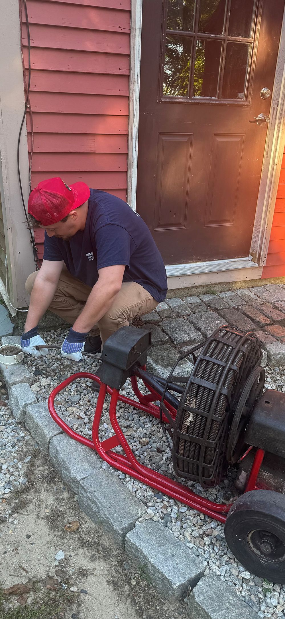 A person in a red hat crouches near a red lawn cart by a building.