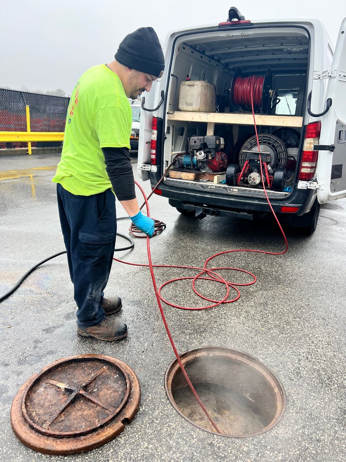 Man in safety vest uses equipment to inspect a manhole on a wet road, van in background.