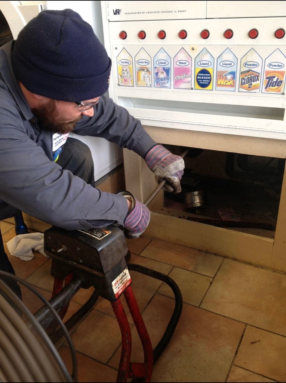 A man in a blue hat uses a drain snake to clear a blockage in a laundry room.