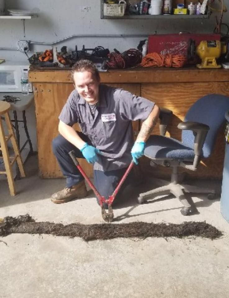 Plumber crouches, wearing gloves, cutting a long hair clog with large shears on a garage floor.
