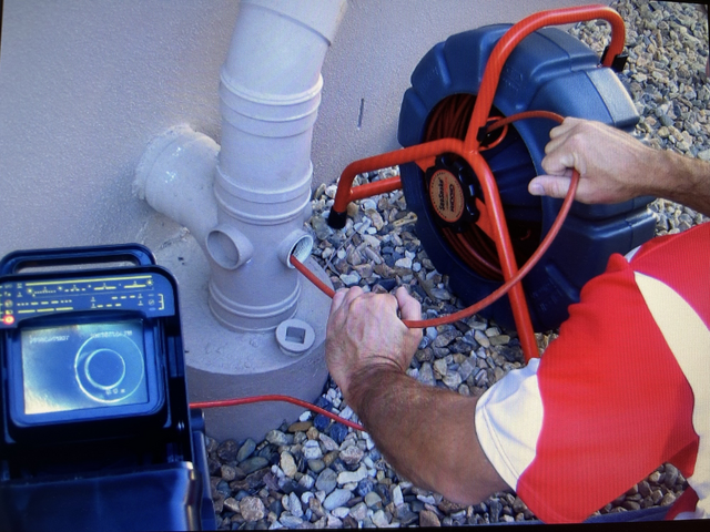 Man using a sewer camera to inspect pipes. Camera monitor, red cable, and reel are visible.
