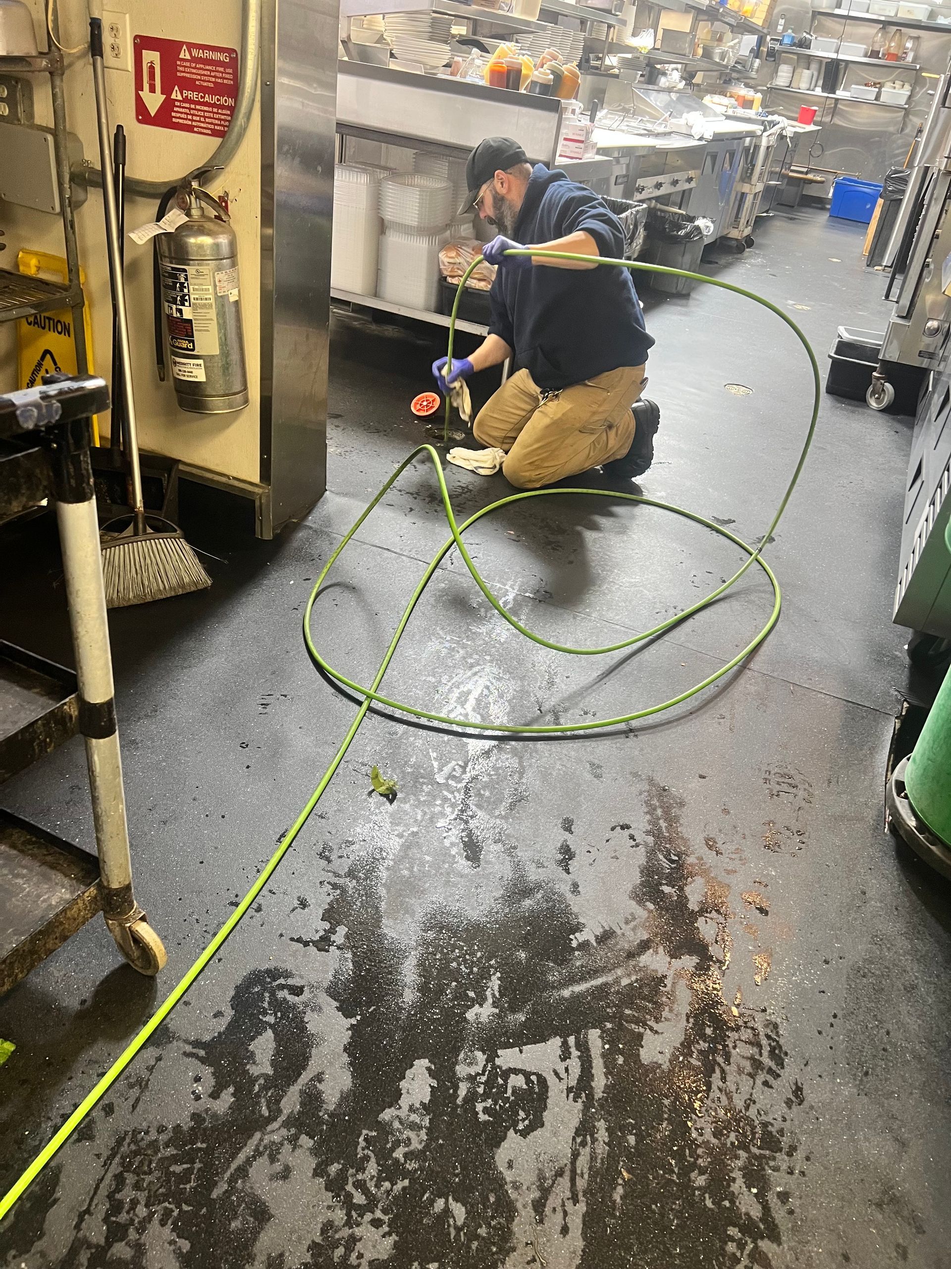 A person kneels on a wet floor, working with a green hose in a commercial kitchen.