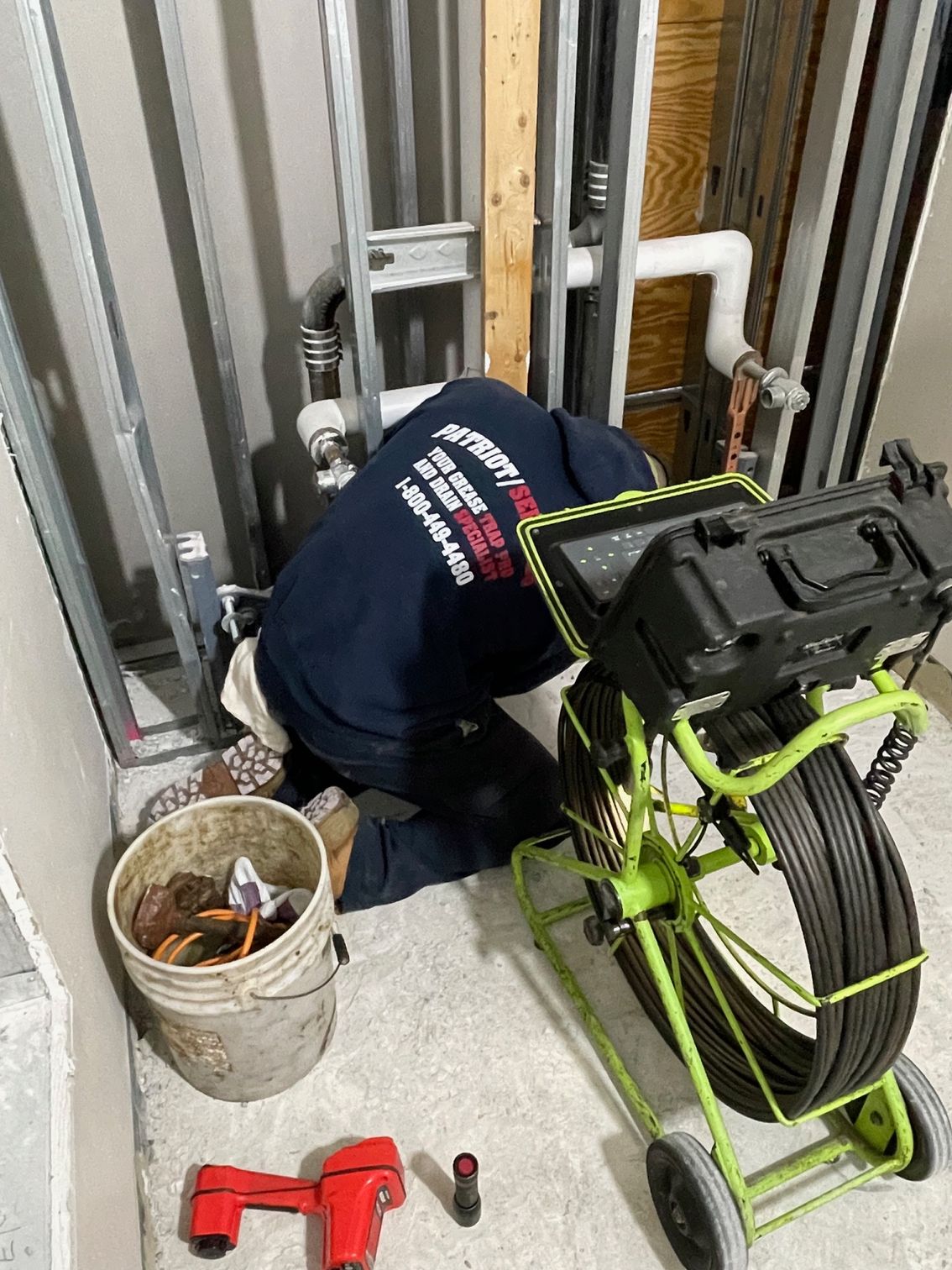 Plumber in a utility room using a sewer camera to inspect pipes. A bucket, and tools are nearby.