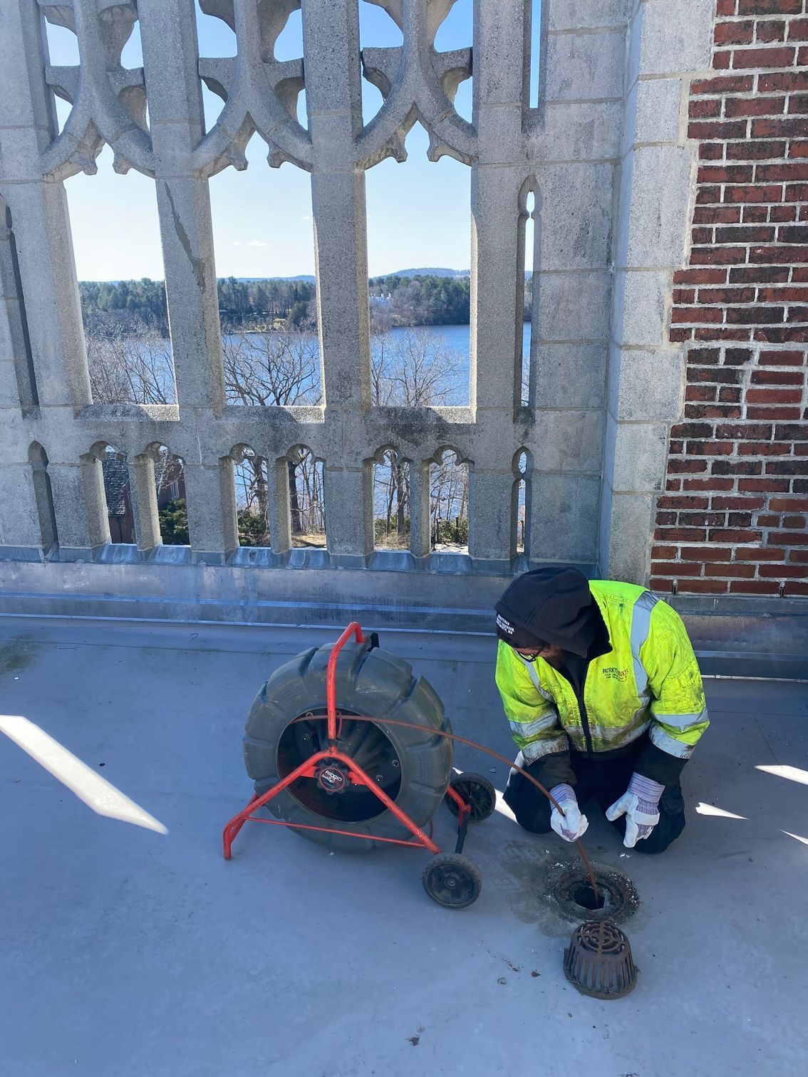 Person in high-vis jacket kneeling by a sewer camera on a rooftop, with a water view behind a stone archway.
