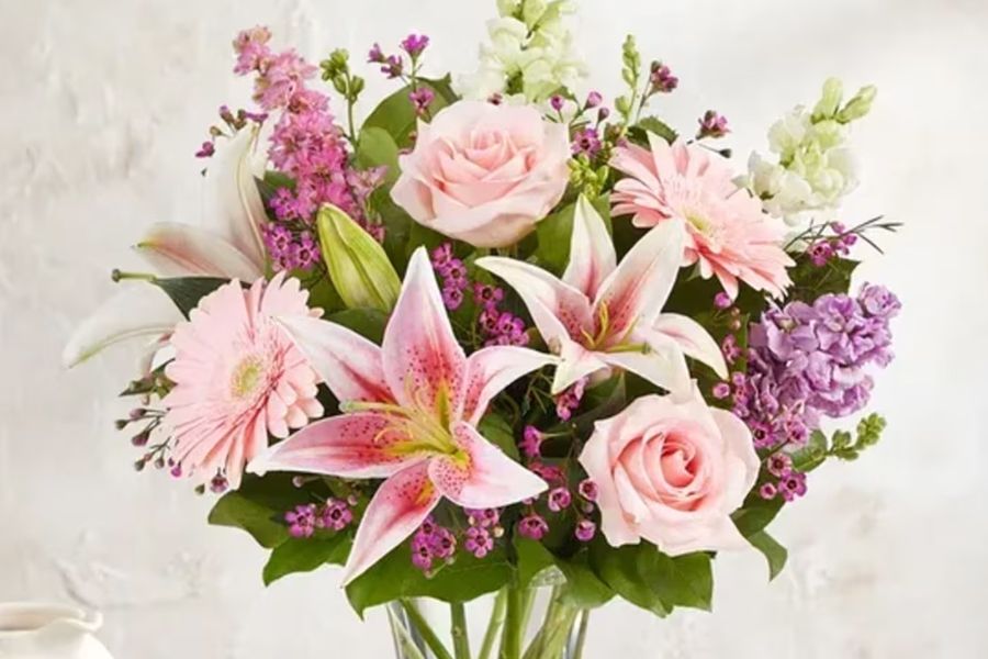 Bouquet of light pink roses, lilies, and daisies, with purple and white accents, in a clear glass vase.