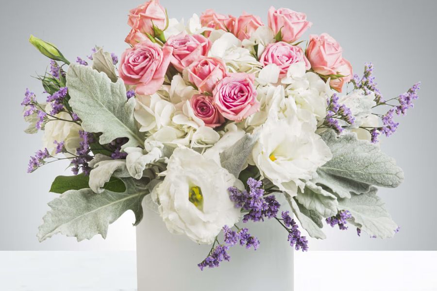 Close-up of a floral arrangement in a white vase. Pink roses, white flowers, and lavender accents are visible.