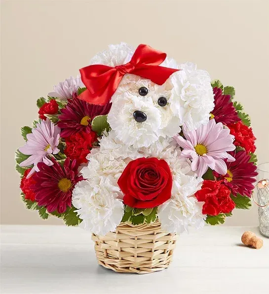 A white dog with a red bow is in a basket of flowers.