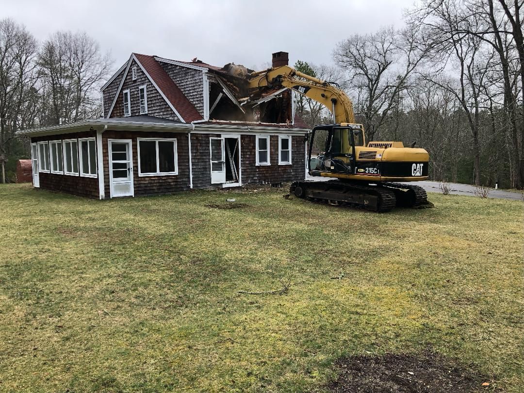 A yellow excavator is demolishing a house in a grassy field.
