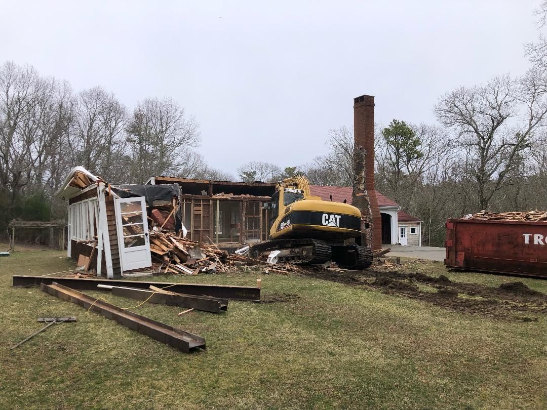A yellow excavator is demolishing a house in a field.