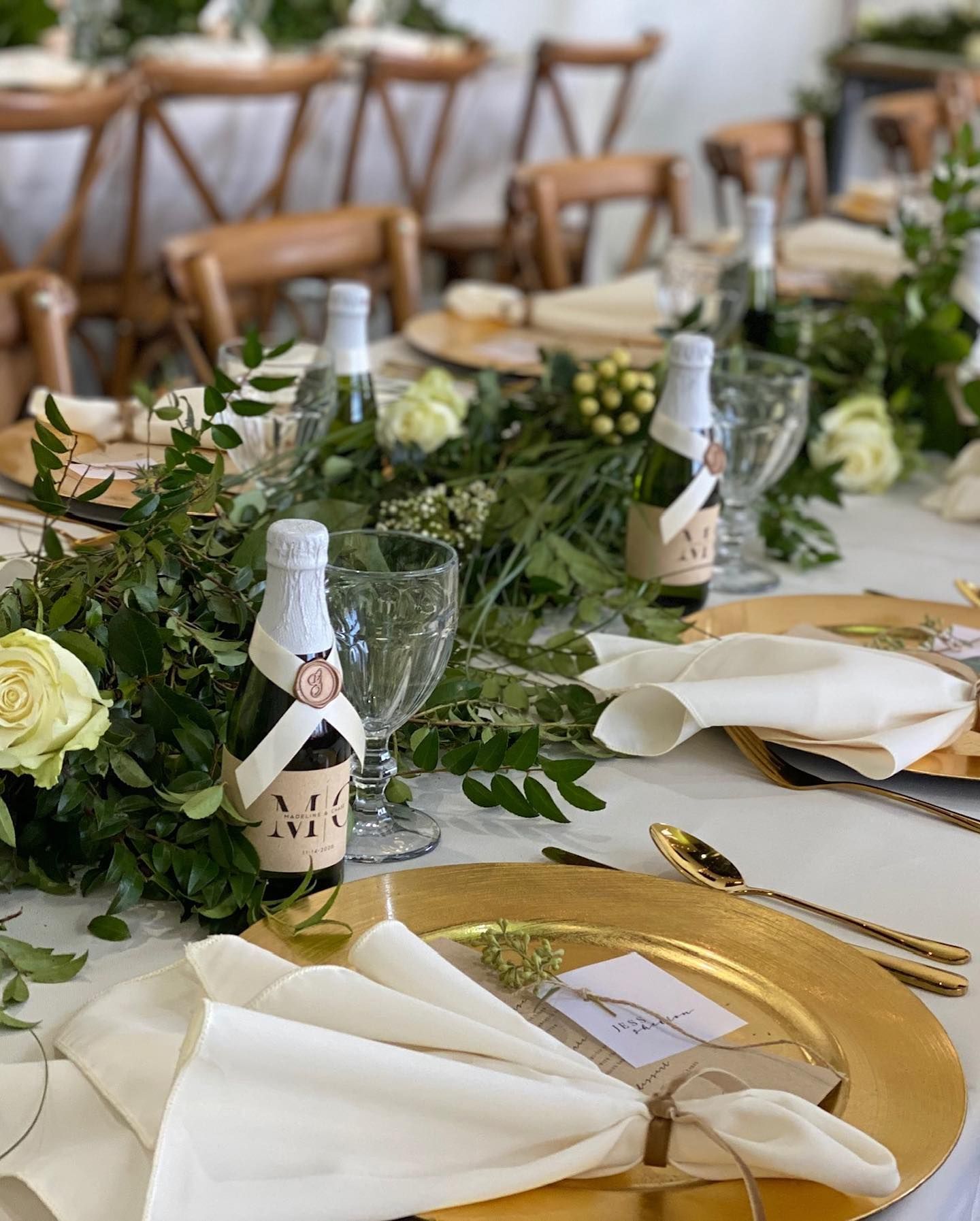 Wedding reception table setting with gold chargers, white napkins, champagne bottles, and greenery.