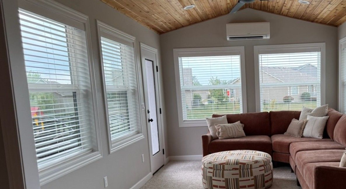 Sunroom with brown sectional sofa, ottoman, and windows with blinds; wood ceiling.
