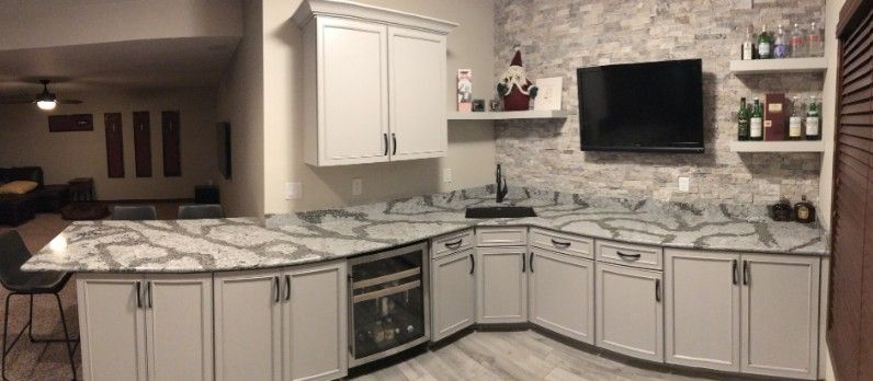 L-shaped wet bar with white cabinets, gray countertops, stone backsplash, and a TV.