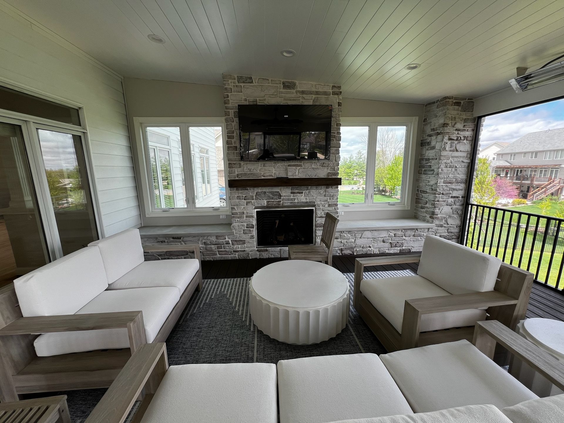 Cozy outdoor living room with fireplace, white couches, rug, and windows overlooking a yard.