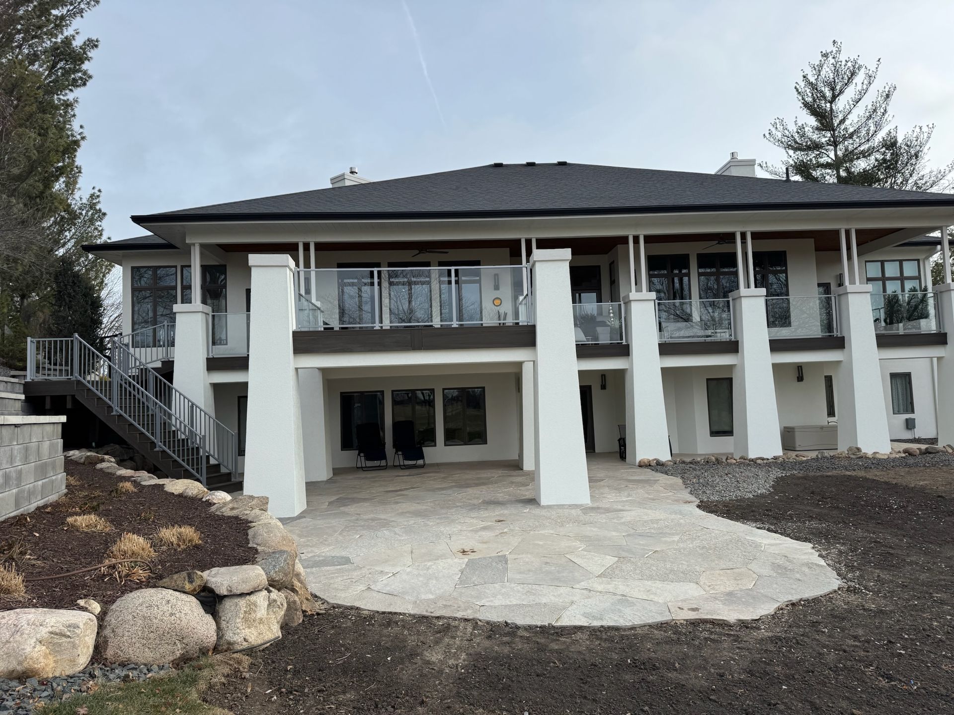 Two-story white house with a black roof and stone patio. Large windows and balcony. Cloudy sky.