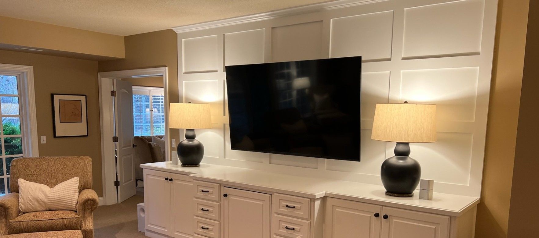 White built-in wall unit with a black TV, two lamps, and cabinets. A chair and doorway are to the left.