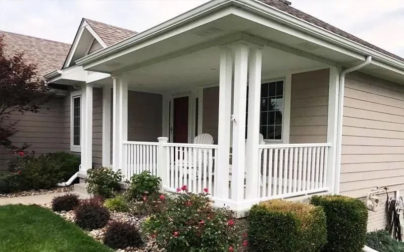 Beige house with a white porch featuring vertical railings, surrounded by green and red bushes.