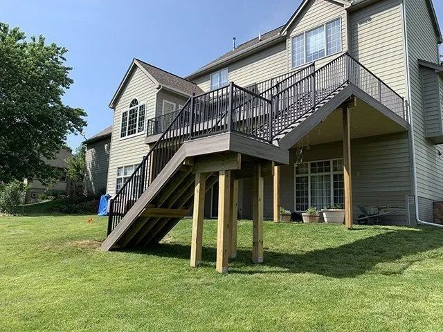 Raised wooden deck and stairs attached to a beige house, with black railings, supported by wooden posts on a grassy lawn.