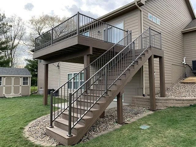 Two-story brown deck with railings and staircase, attached to a light brown house with green lawn.