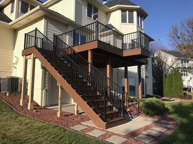 Brown deck with black railing and stairs attached to a beige house, with landscaping.