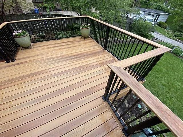 Wooden deck with black railings and two green potted plants. Overlooks a yard with trees and a house.