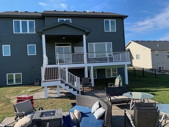 Back of a two-story blue house with a deck and patio furniture in a yard on a sunny day.