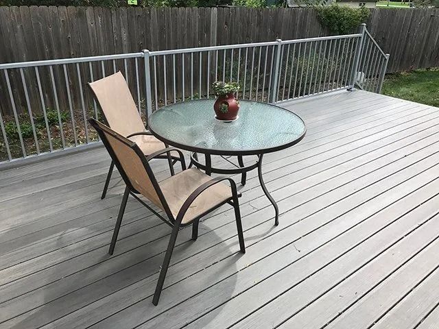 A small outdoor deck with a round glass table, two chairs, and a decorative vase. Gray railing and deck planks.